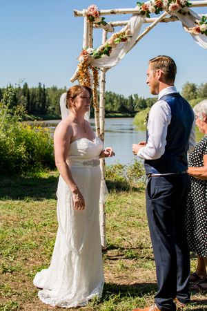 Outdoor wedding ceremony by a river with a rustic wooden arch decorated with coral and peach flowers.