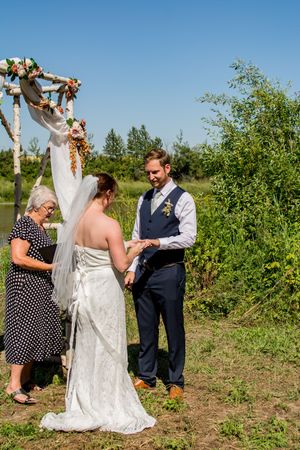 An outdoor wedding ceremony takes place under a floral arch beside a lake on a sunny day.