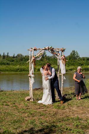 Outdoor wedding ceremony by riverside with wooden arch and gathered guests.