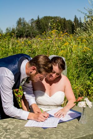 A couple signs their marriage documents outdoors on a sunny day surrounded by wildflowers.