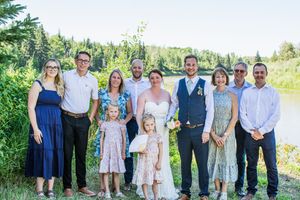 Wedding party group photo with guests in blue and floral dresses posed outdoors near trees.