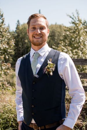 Groom in a navy vest and white shirt smiles while standing in an outdoor garden setting.