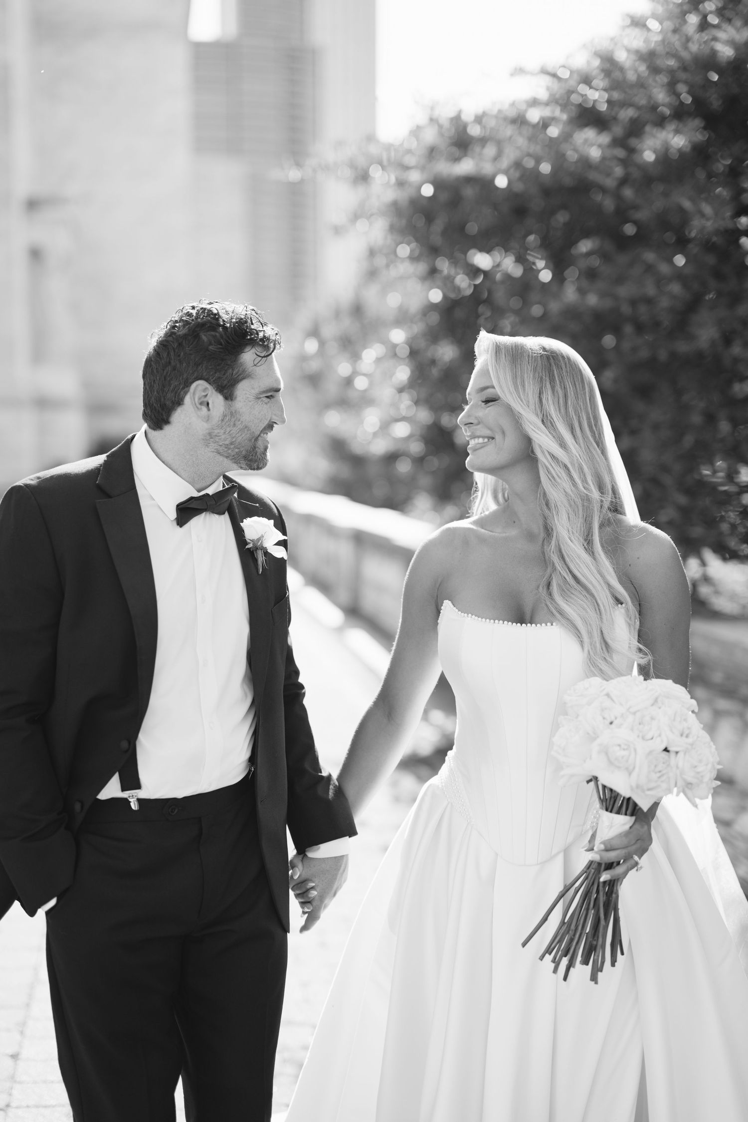 A bride in a strapless white gown holds a bouquet while walking with her groom in a black tuxedo on a sunny day.