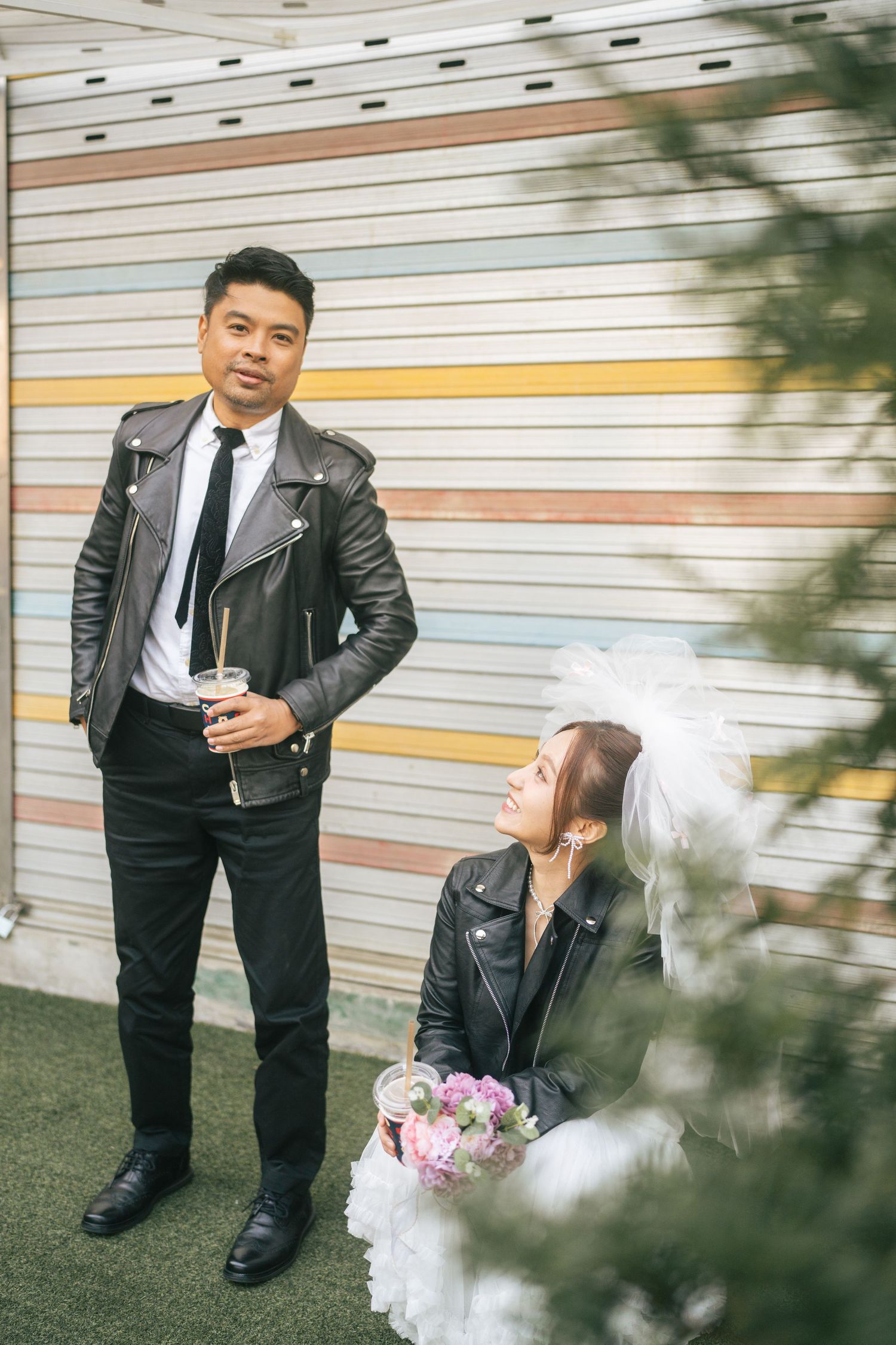 A stylish wedding couple in leather jackets poses against a striped wall with the bride holding a pink bouquet.