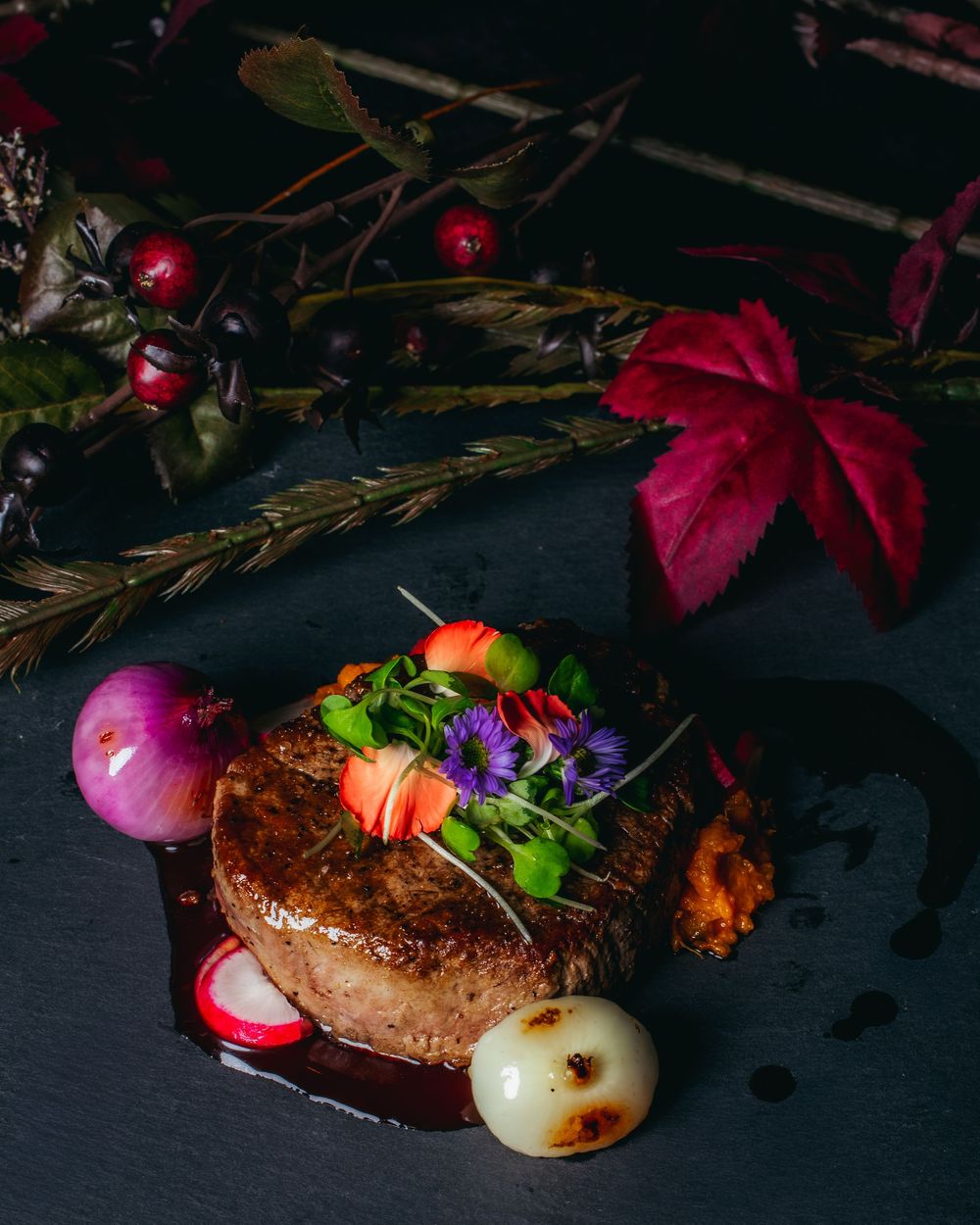 Dark moody food photography of rustic bread loaves with red flowers and autumn leaves on black background.