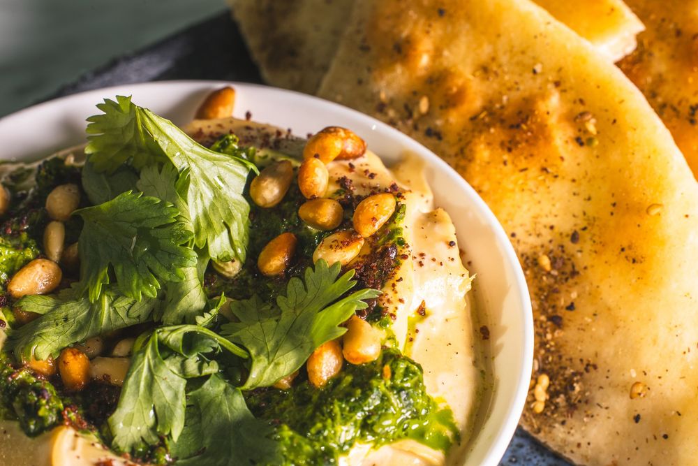 Close up of hummus topped with chickpeas, cilantro and spices served with flatbread on white plate.