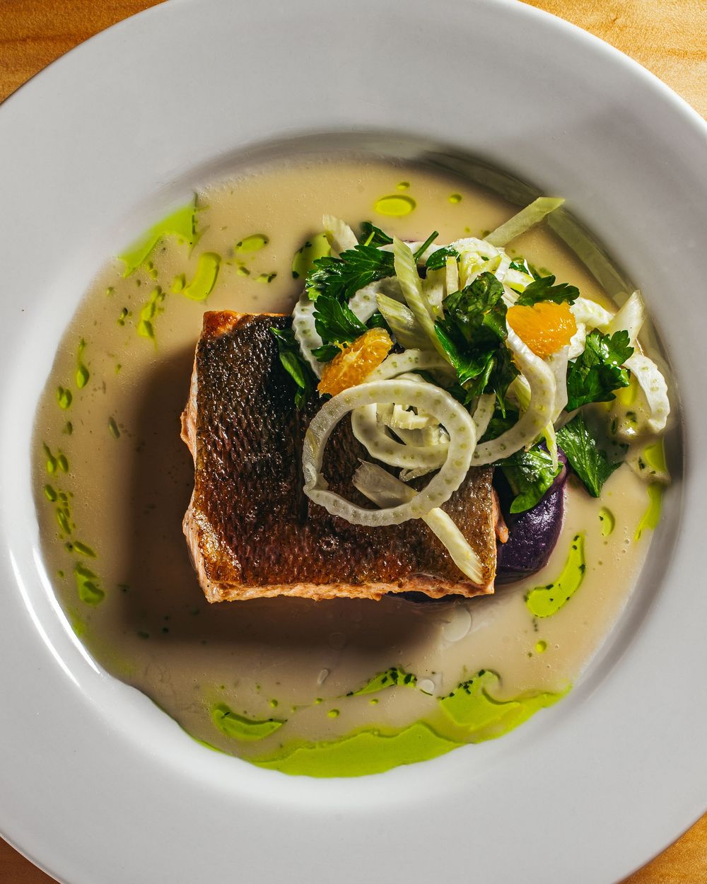 Pan-seared fish fillet served in a light broth with sliced onions, broccoli and herbs on a white plate with green oil drizzle.