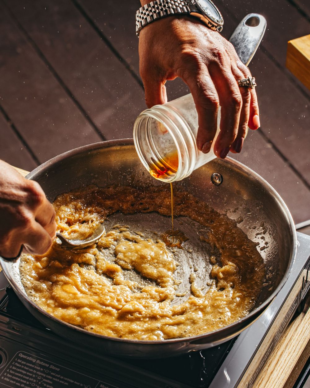 Hands pouring honey or syrup into a sizzling pan with melted butter and caramelized ingredients.