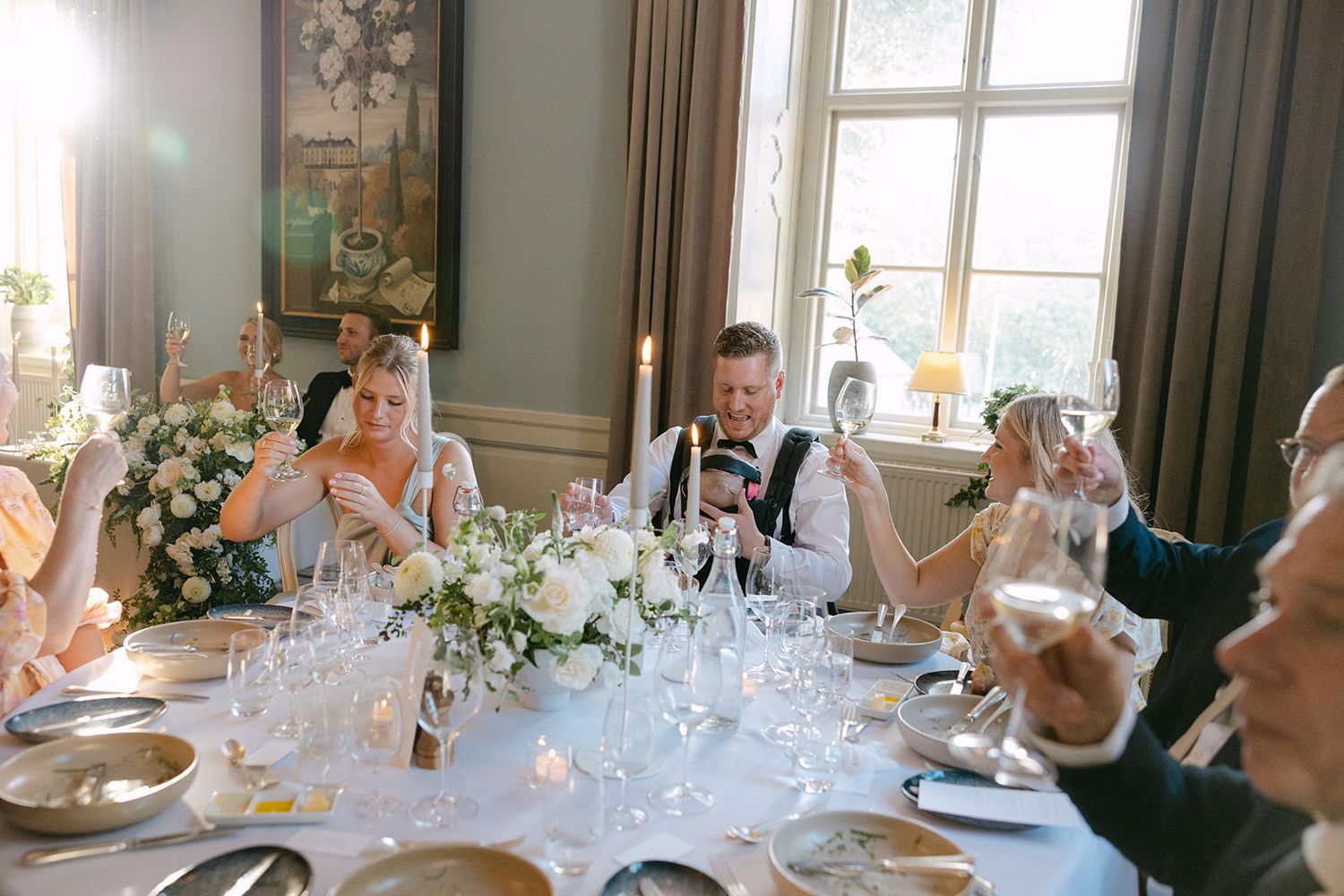 Elegant wedding reception tables set with white linens, floral centerpieces and glassware in a bright sunlit dining room.
