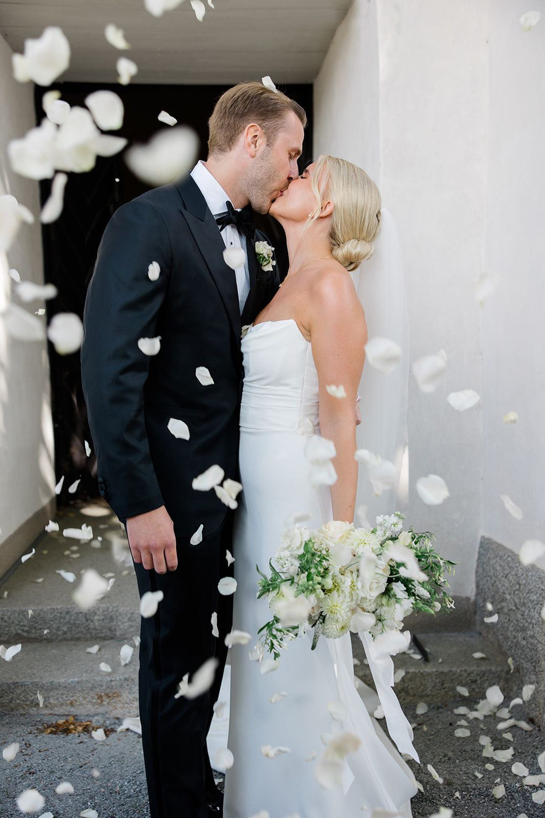 A bride and groom share a romantic kiss as white rose petals fall around them during their wedding celebration.
