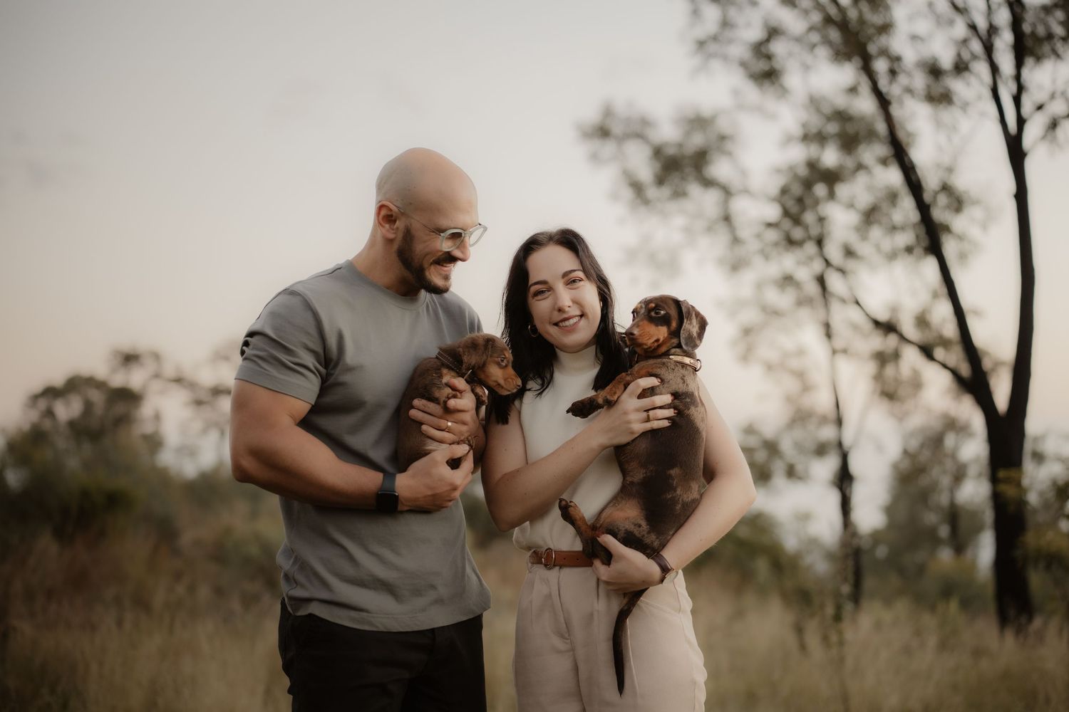 A couple in casual attire holds two small puppies while standing outdoors in a natural setting at sunset.