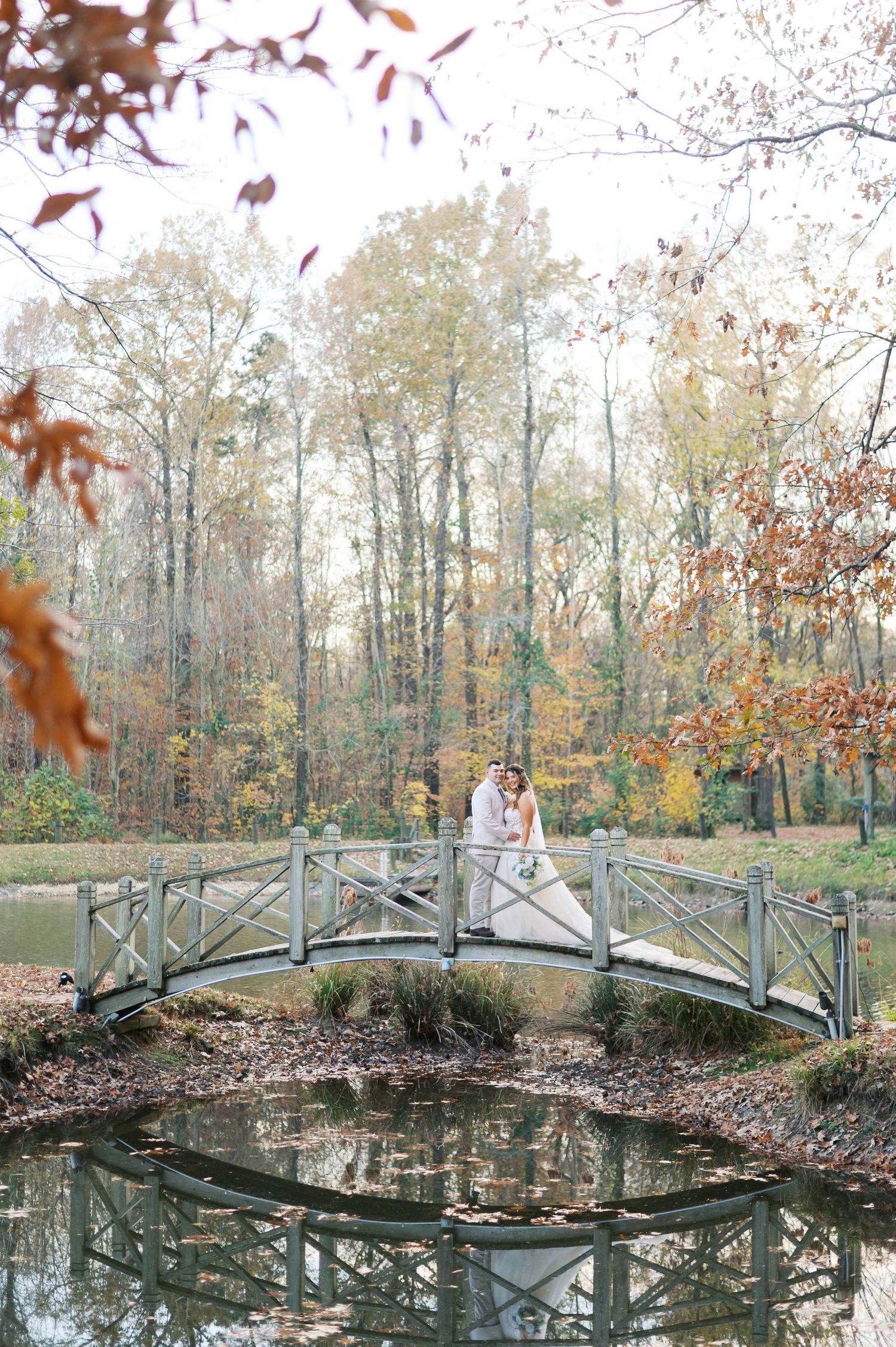 A rustic wooden footbridge spans over a tranquil pond surrounded by autumn trees with falling leaves.