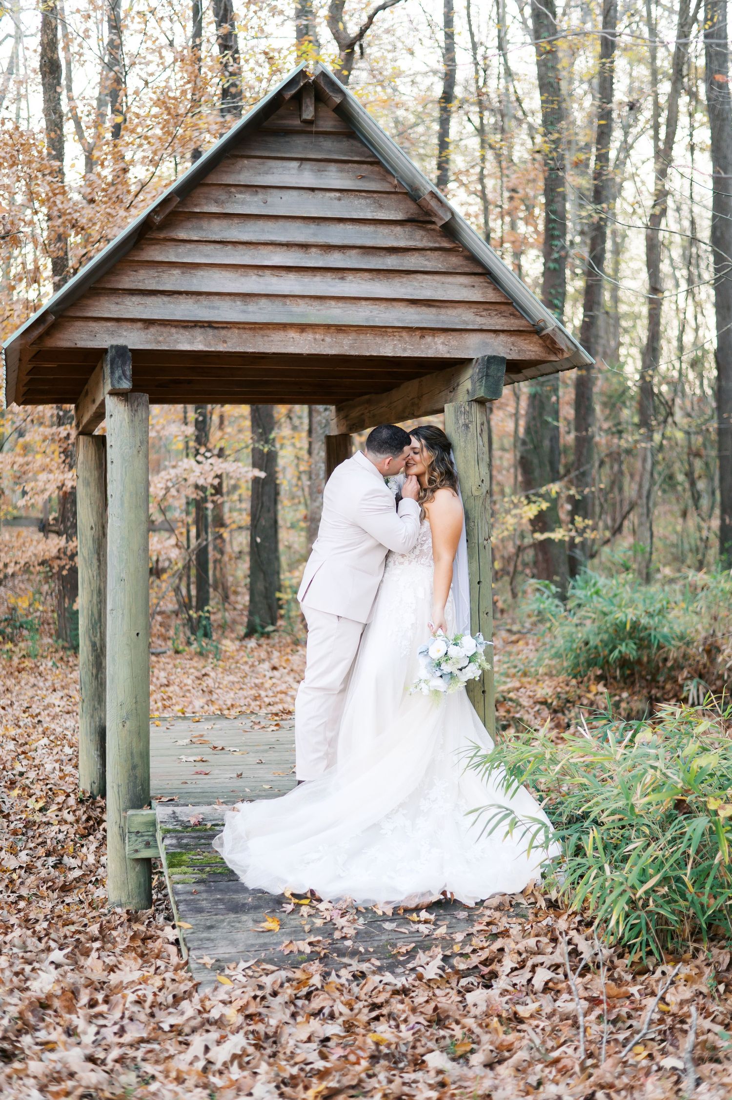 Romantic wedding couple shares intimate moments under rustic wooden gazebo in autumn forest setting.