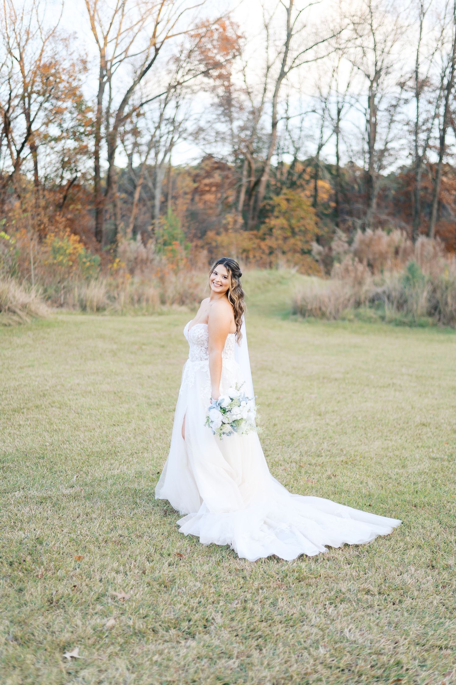 A bride in a flowing white gown sits gracefully in a rustic autumn field surrounded by fall foliage.