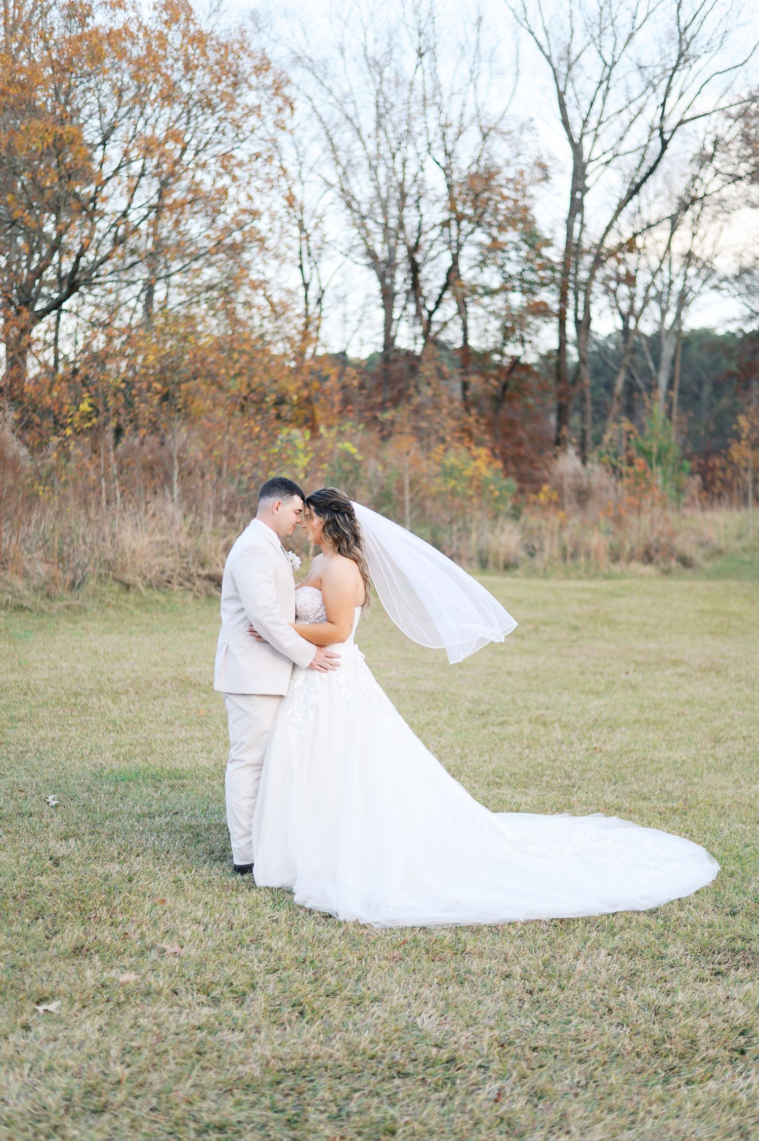 A romantic wedding couple embraces in a field with autumn trees in the background during their outdoor photo session.