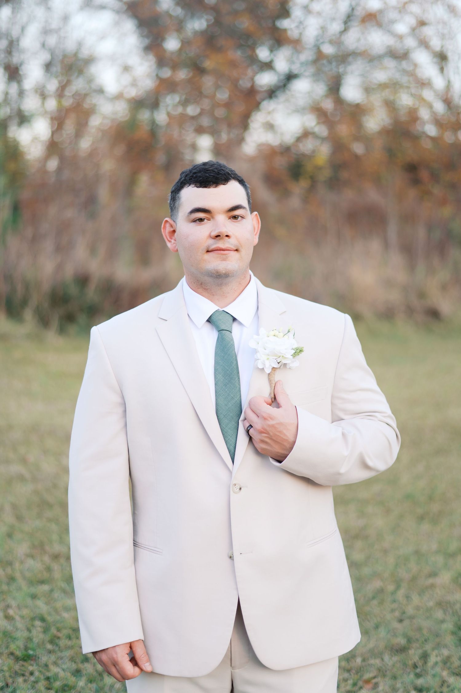 A groom in a cream-colored suit and sage green tie poses for formal wedding portraits outdoors.
