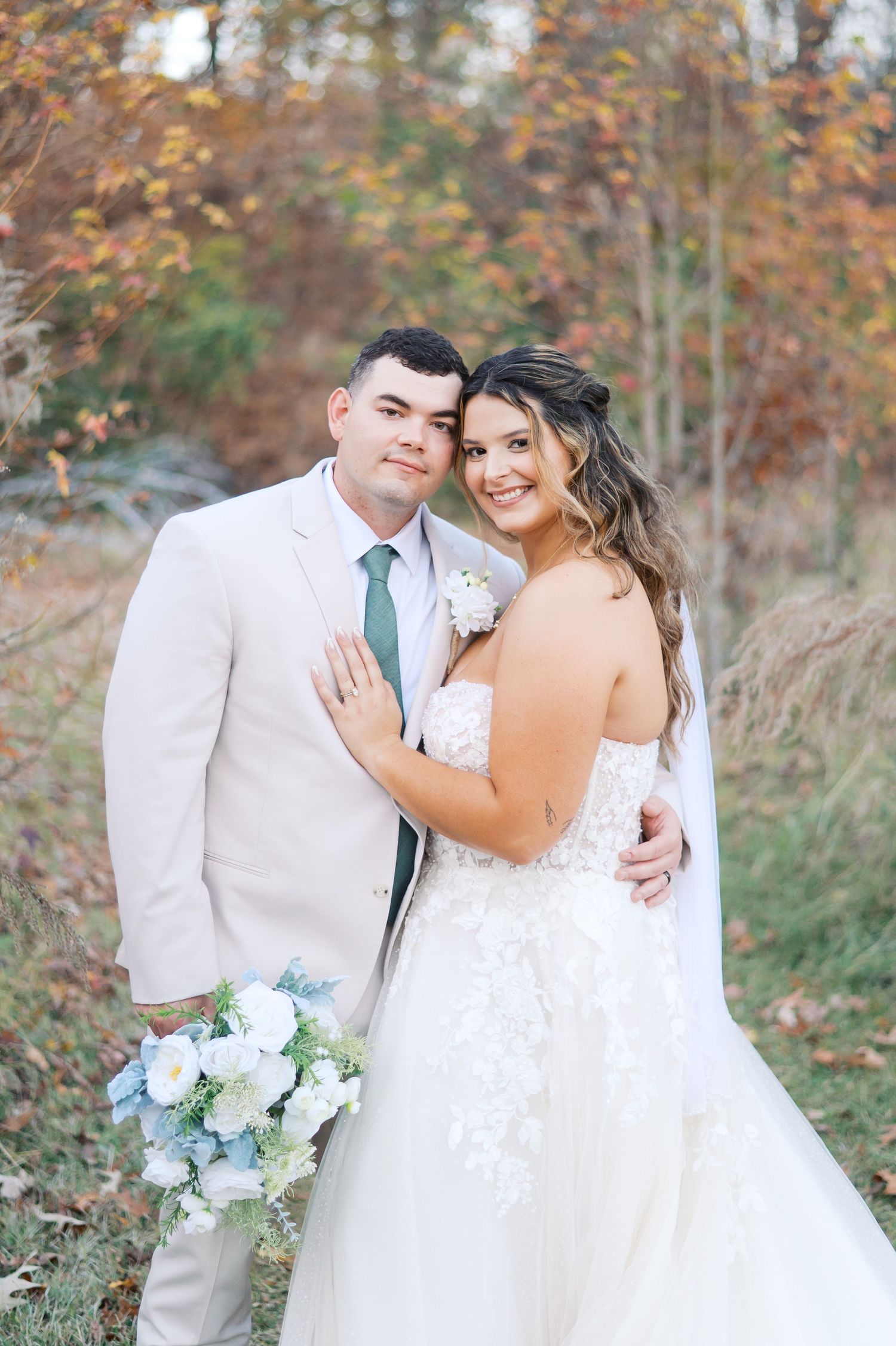 A romantic autumn wedding portrait features a couple in white attire with a soft blue and white bouquet against fall foliage.