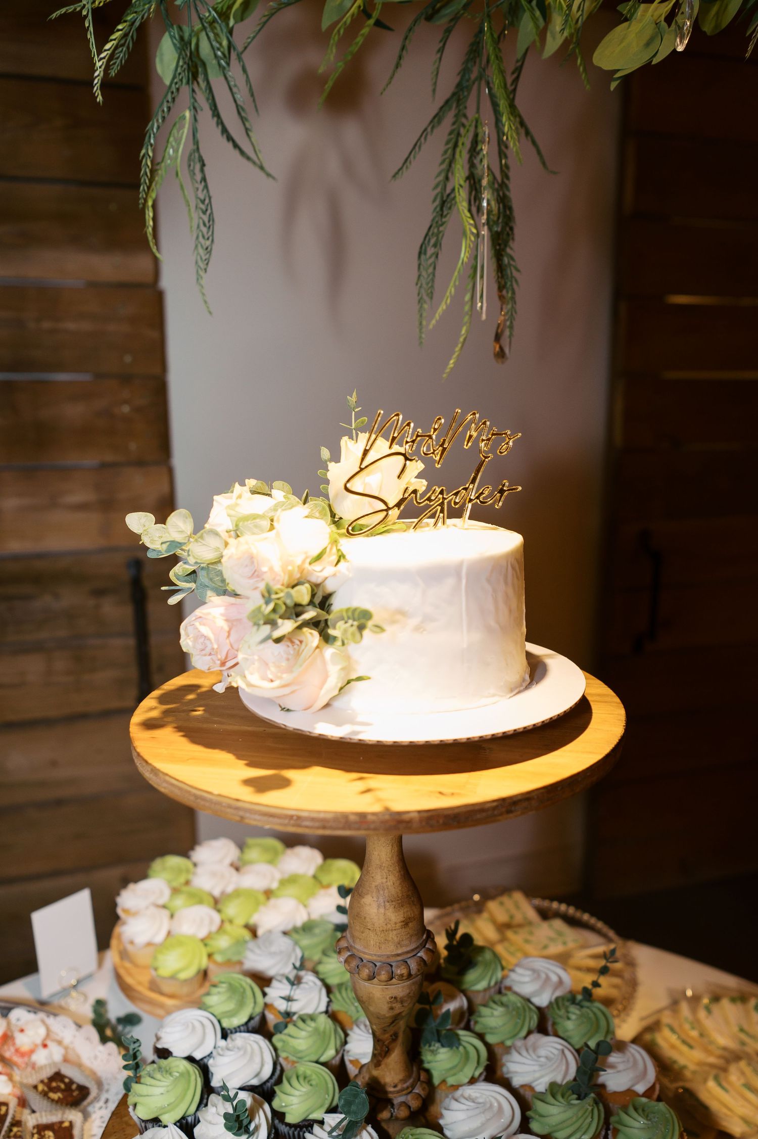 White wedding cake on gold stand surrounded by delicate floral arrangements and greenery in rustic venue setting.
