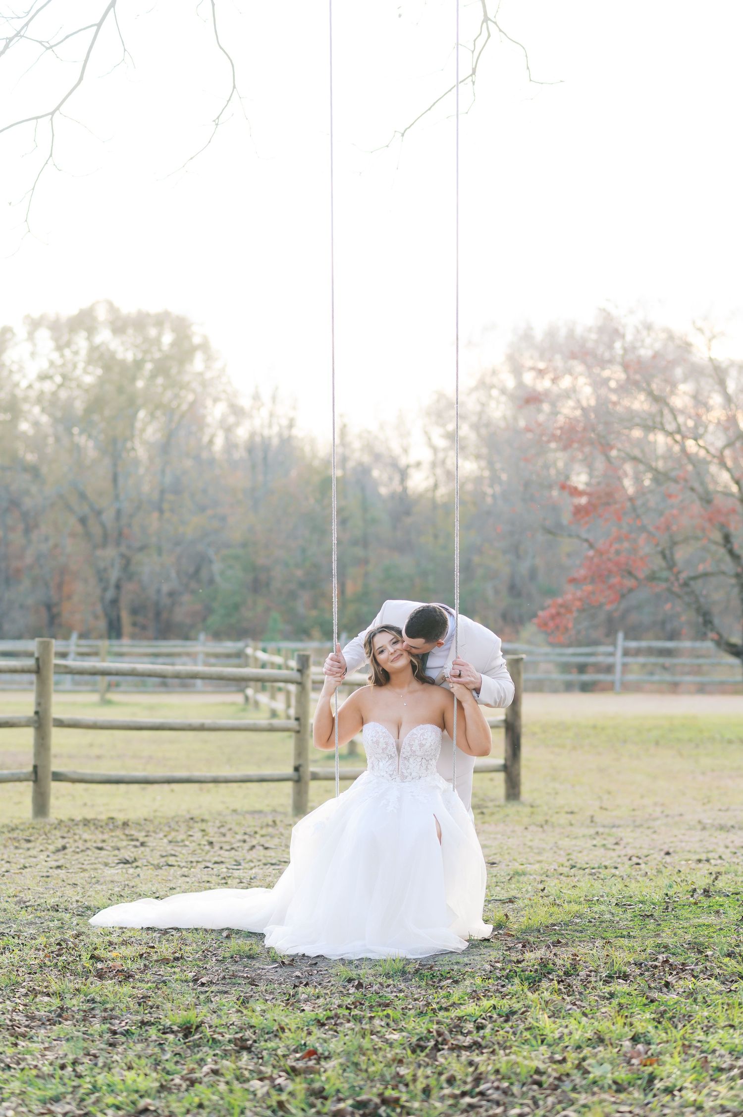 A wedding couple poses playfully on the grass in front of a rustic wooden fence during an autumn photoshoot.