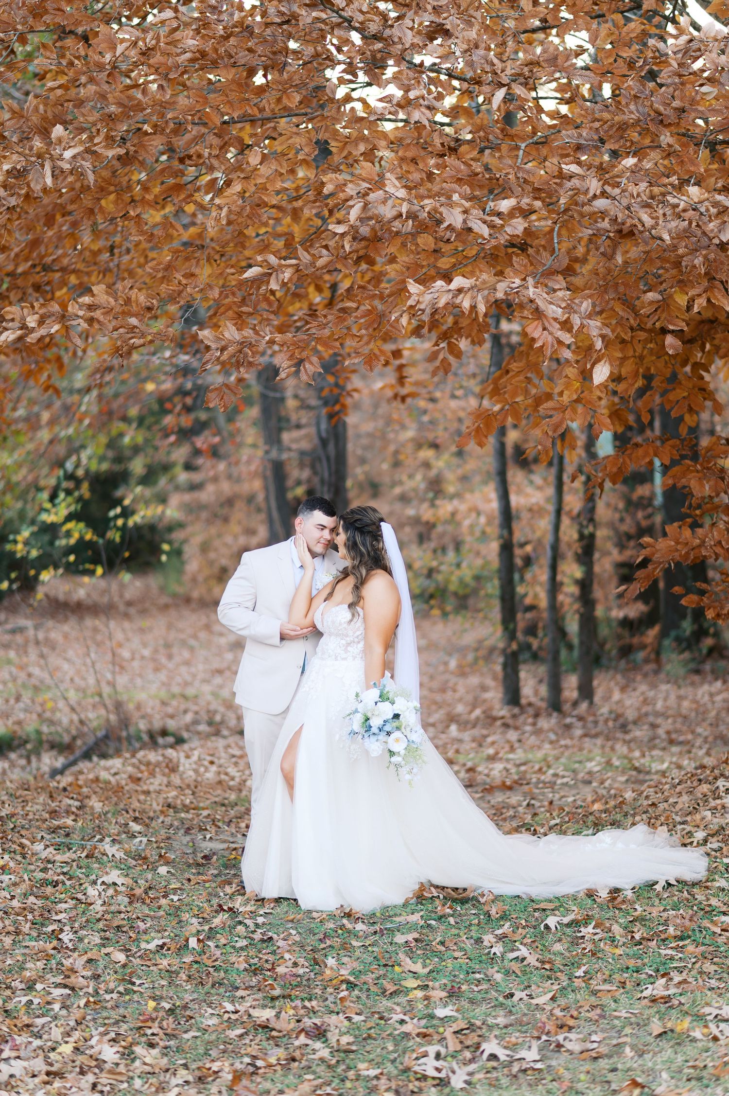 A romantic autumn wedding photo with a couple embracing under orange fall foliage in a wooded setting.