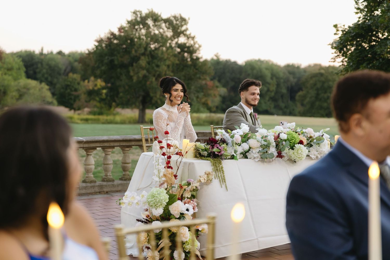 Elegant outdoor wedding reception setup with rustic wooden tables and floral centerpieces beside a tranquil lake at sunset.