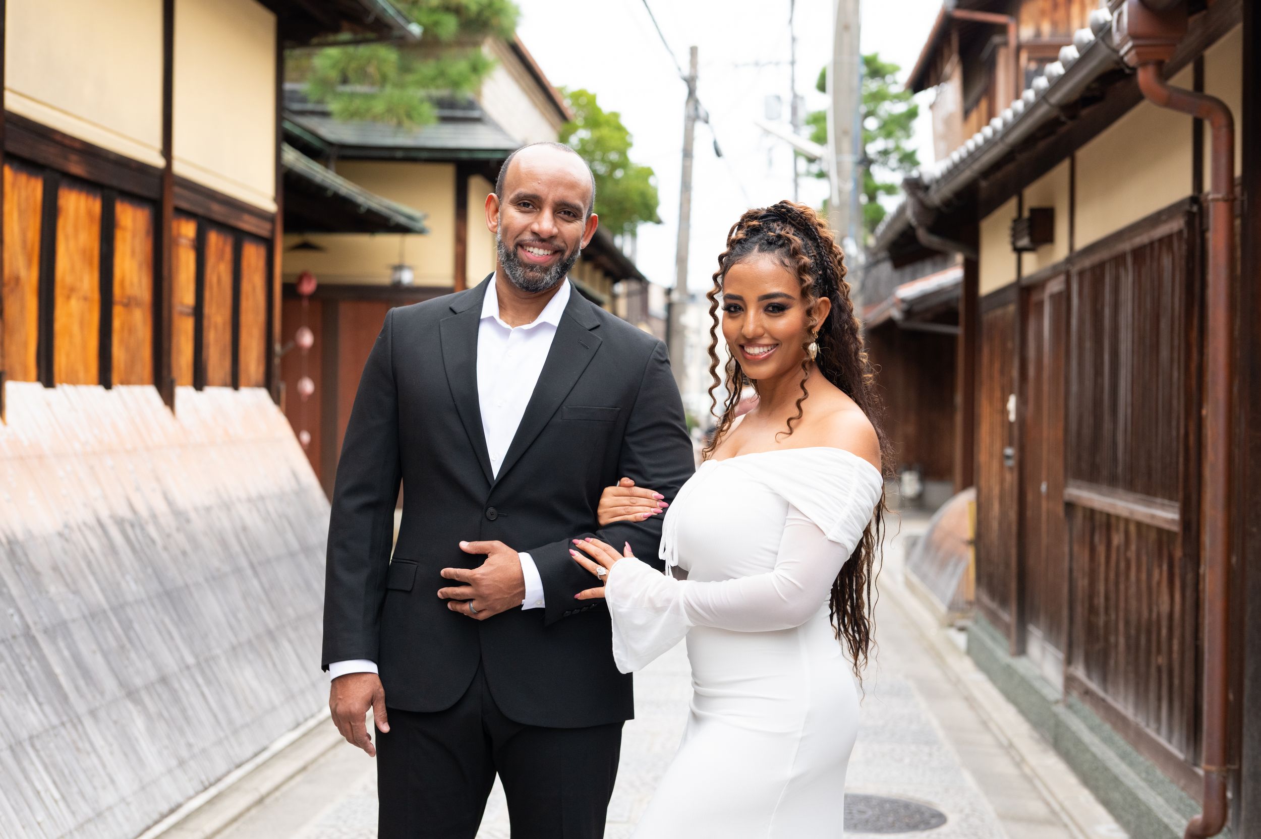 Couple poses in Kyoto alley, woman in white off-shoulder gown, man in black suit, traditional wooden buildings.