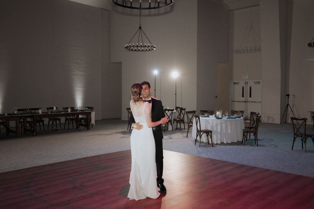A couple shares their first dance in an elegant white ballroom with chandelier lighting overhead.