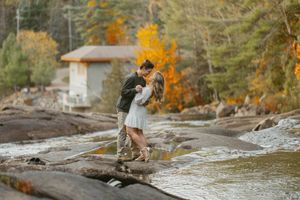 A couple embracing on rocky riverbank during autumn with golden trees in background.