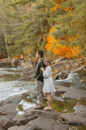 A couple standing on rocks by a river surrounded by fall foliage.