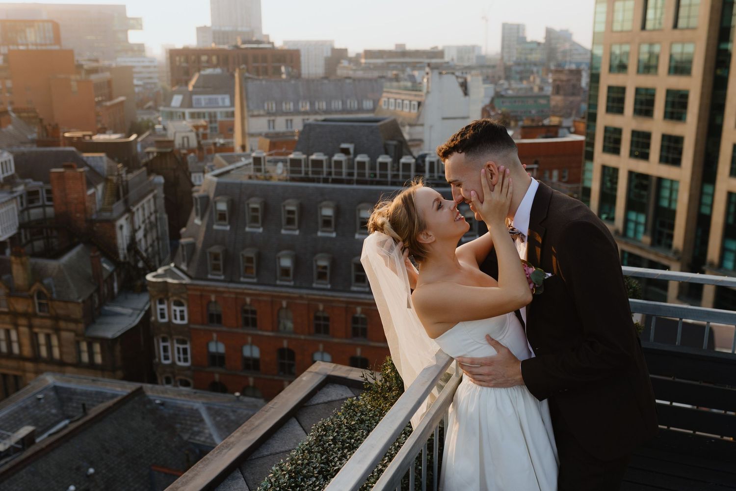 A couple embraces on a rooftop terrace overlooking a cityscape at sunset with brick buildings in the background.