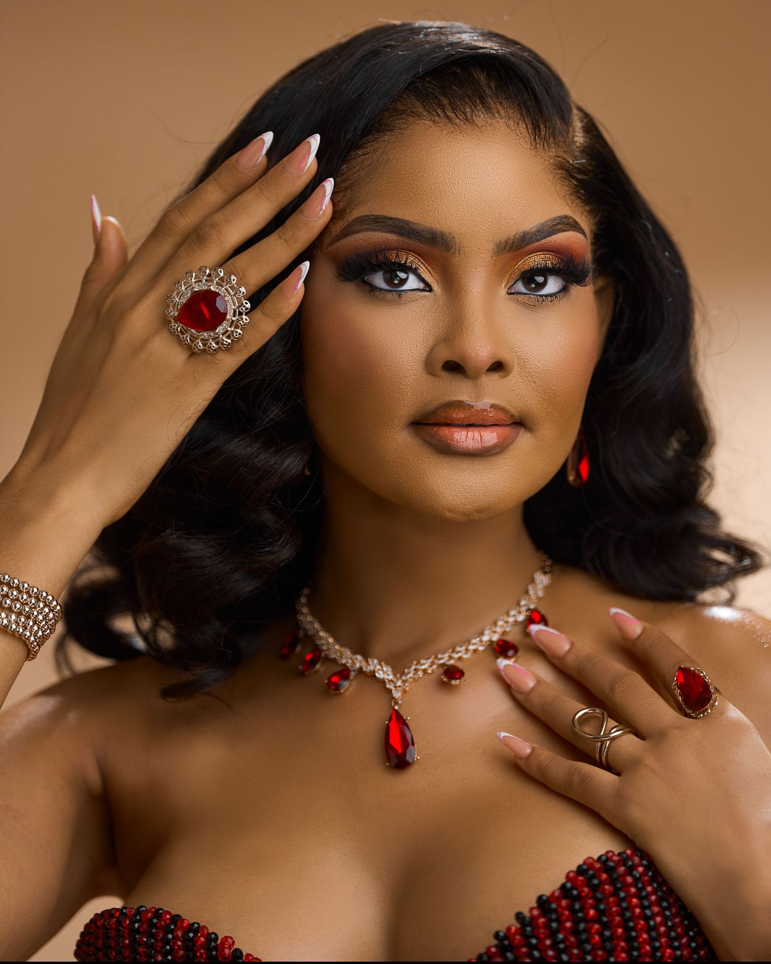 A glamorous Black woman in red and black jewelry poses elegantly against a warm beige background.