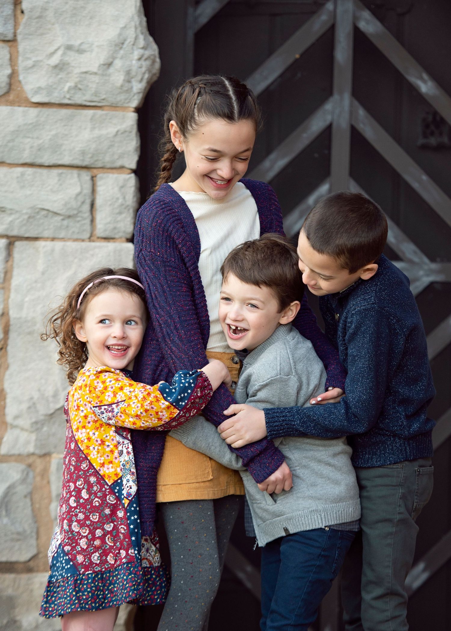 Four happy children hugging and laughing together outdoors near a stone wall and wooden door.