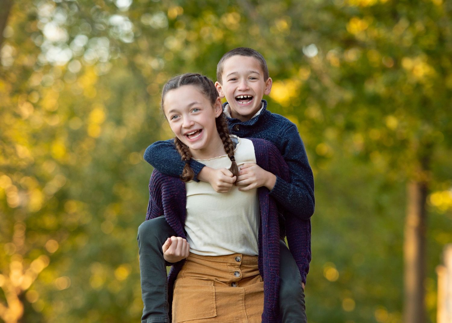 Two laughing children play outdoors in autumn, girl giving piggyback ride to boy among golden trees.