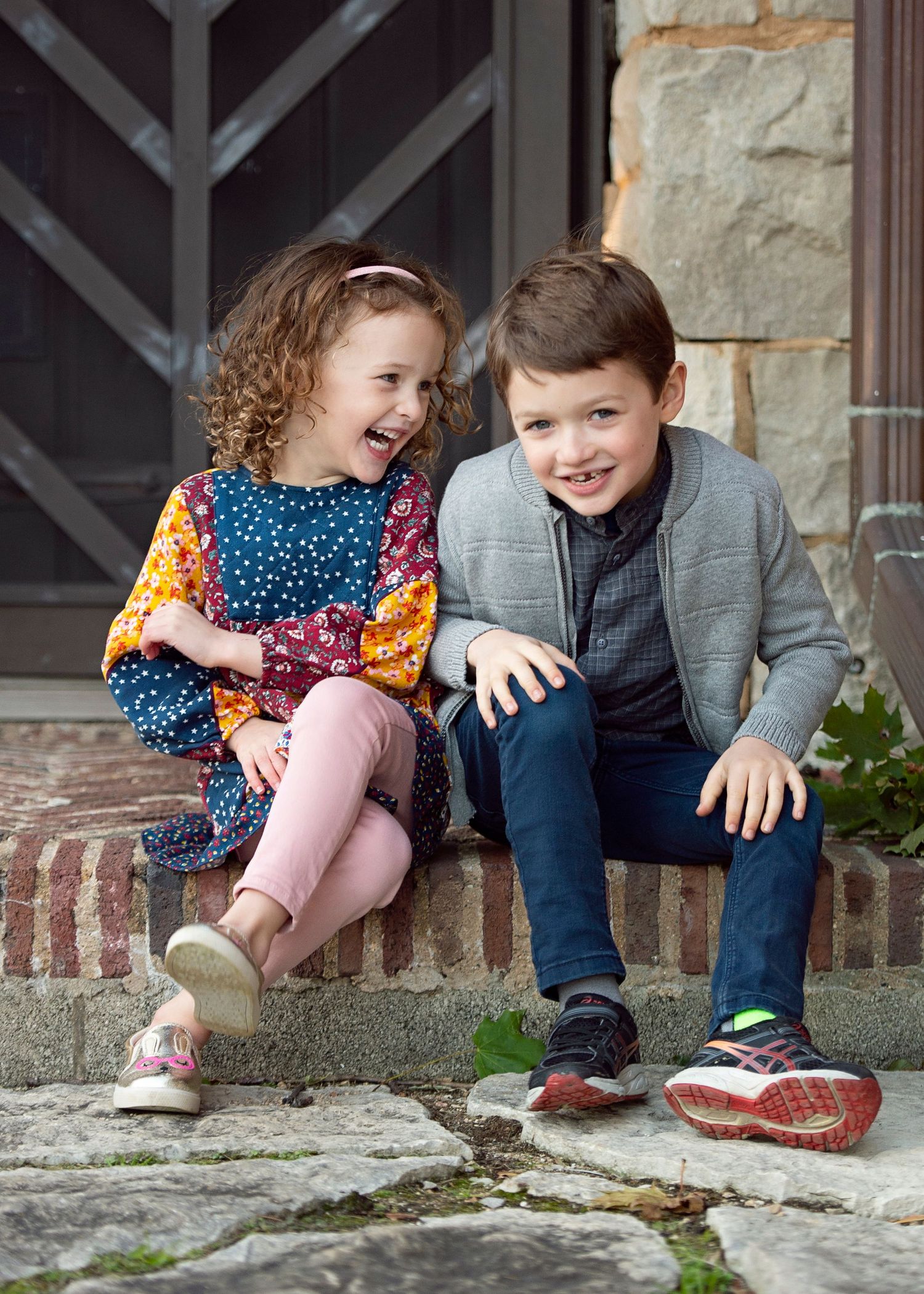 Two young children sitting on stone steps laughing together, girl in colorful dress and boy in gray jacket and jeans.