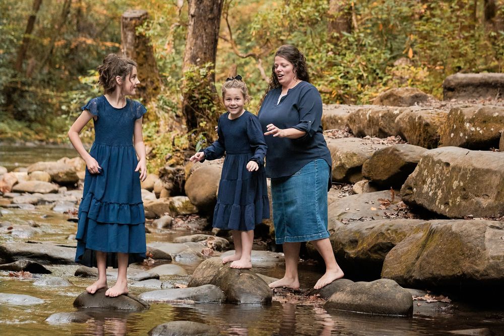 Family wearing coordinated blue outfits sits together on large rocks by a peaceful stream in autumn.