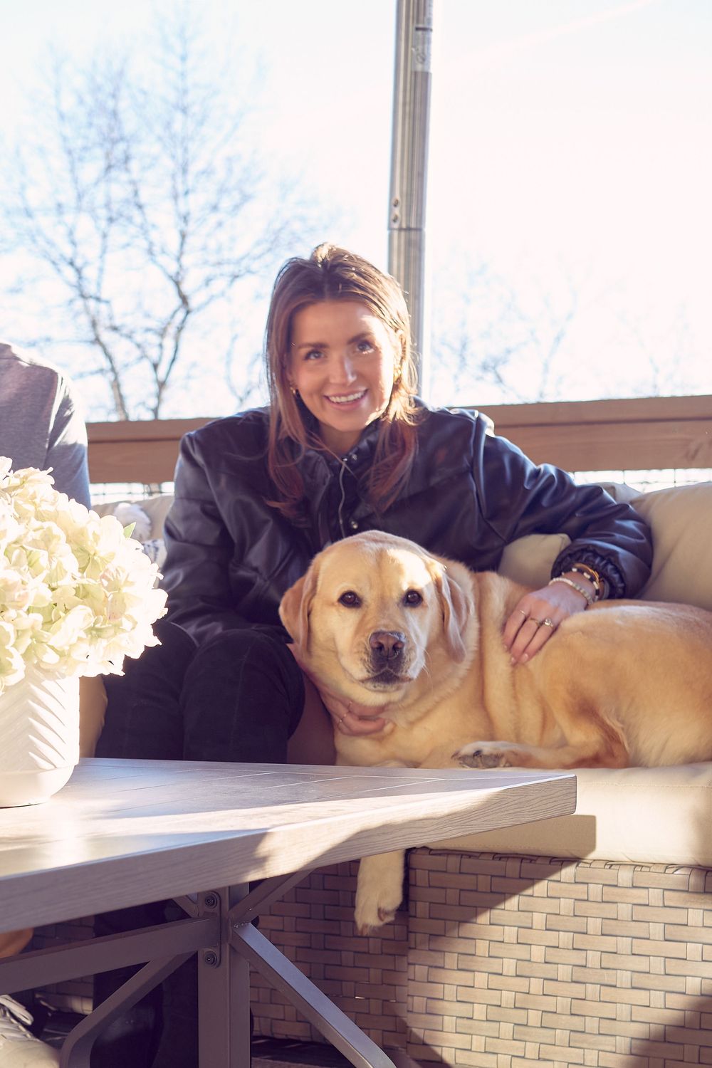 A yellow Labrador retriever lounges on an outdoor patio couch next to someone in a black jacket on a sunny day.