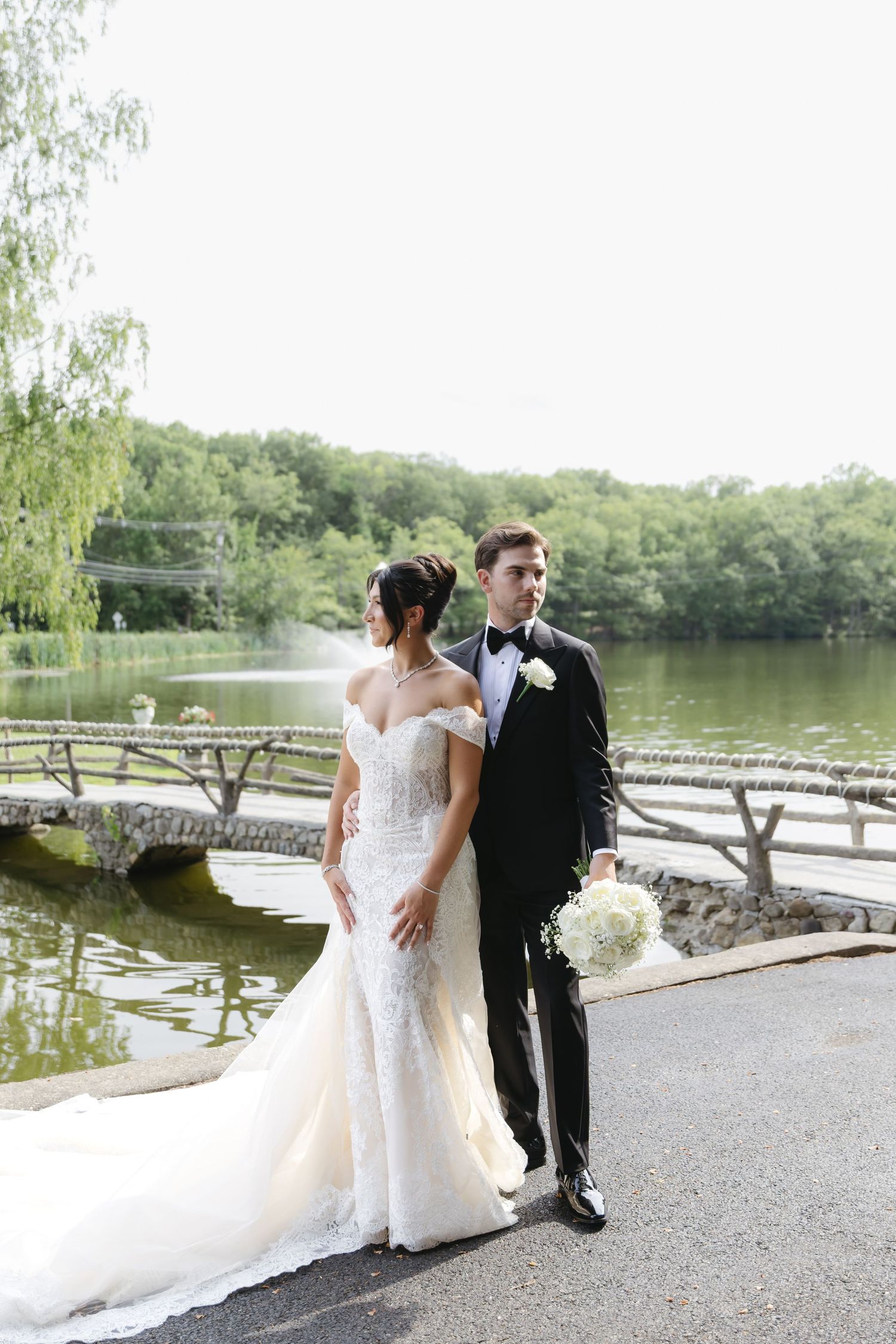 A romantic wedding couple poses by a serene lake with a stone bridge in the background on their special day.
