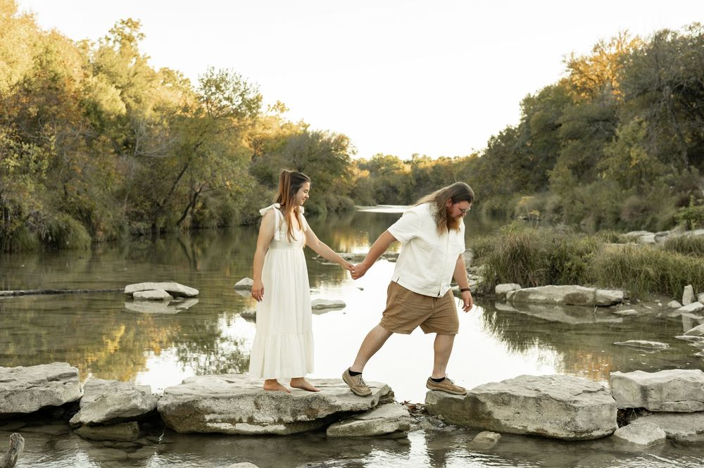 A couple in white outfits poses together on rocks by a serene lake during golden hour sunset.