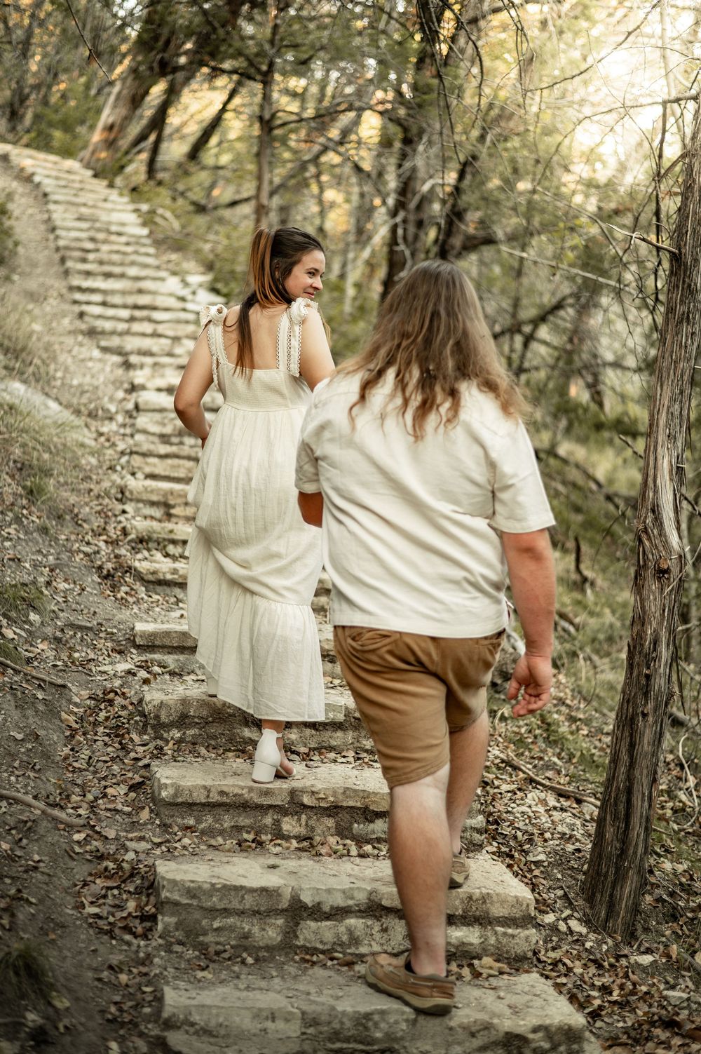 Two people in white and brown outfits walking up stone steps in a wooded area during golden hour.