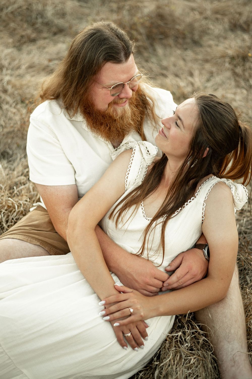A romantic couple in white clothing embraces tenderly while sitting in a rustic outdoor setting.