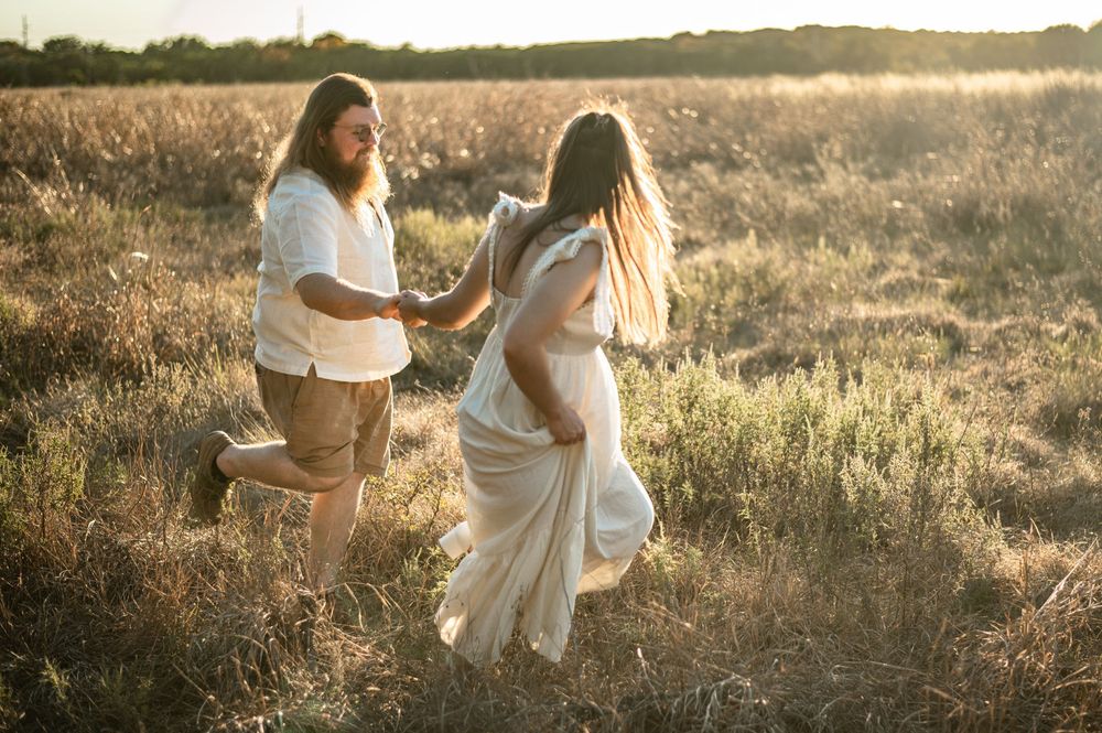 A couple embraces in a sunlit meadow during golden hour, wearing white flowy outfits against tall grass.