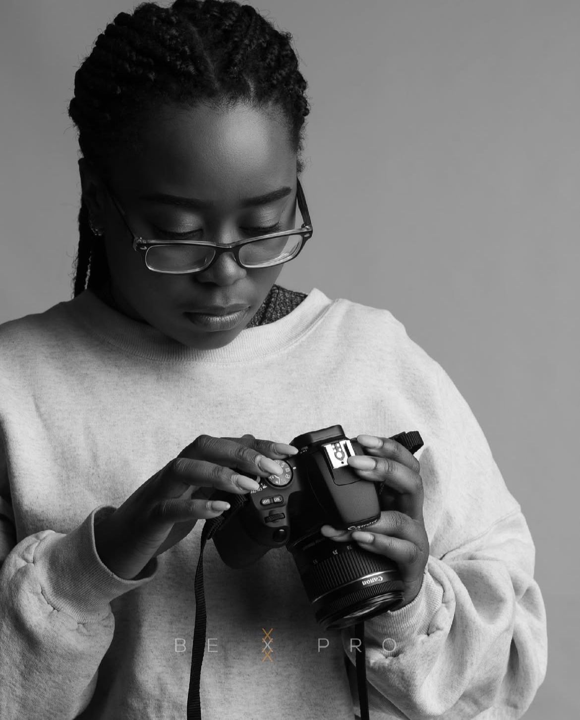 Black and white portrait of a young Black woman with braids and glasses examining a DSLR camera.