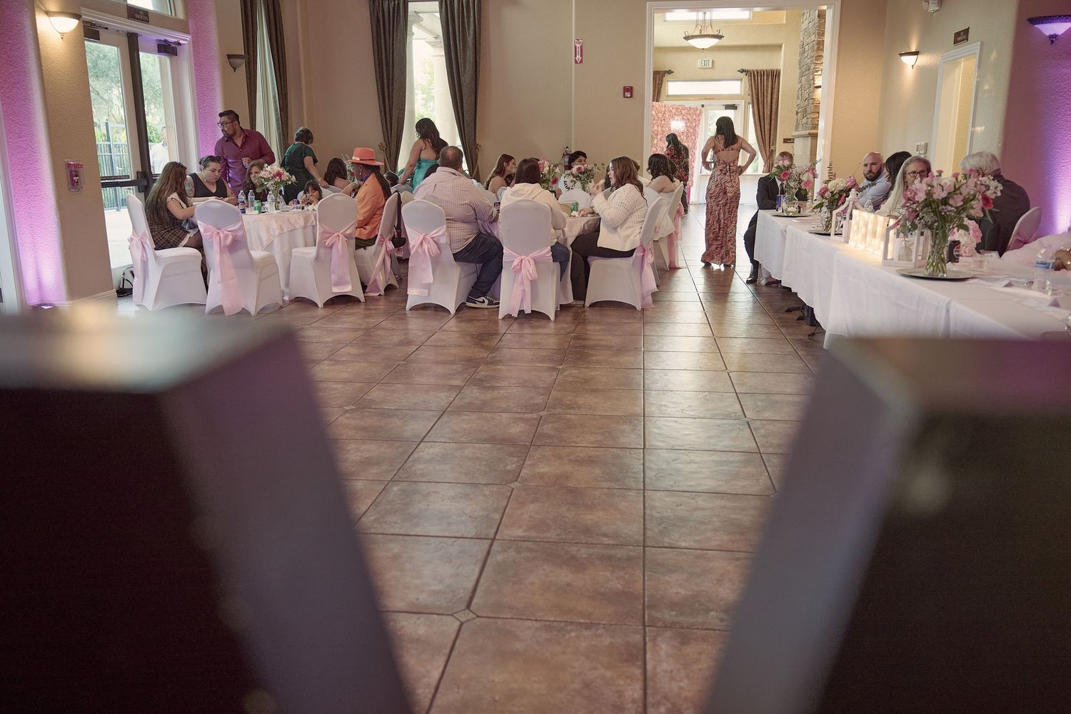 A large wedding reception hall with white table linens, purple accent lighting and a tiled dance floor.