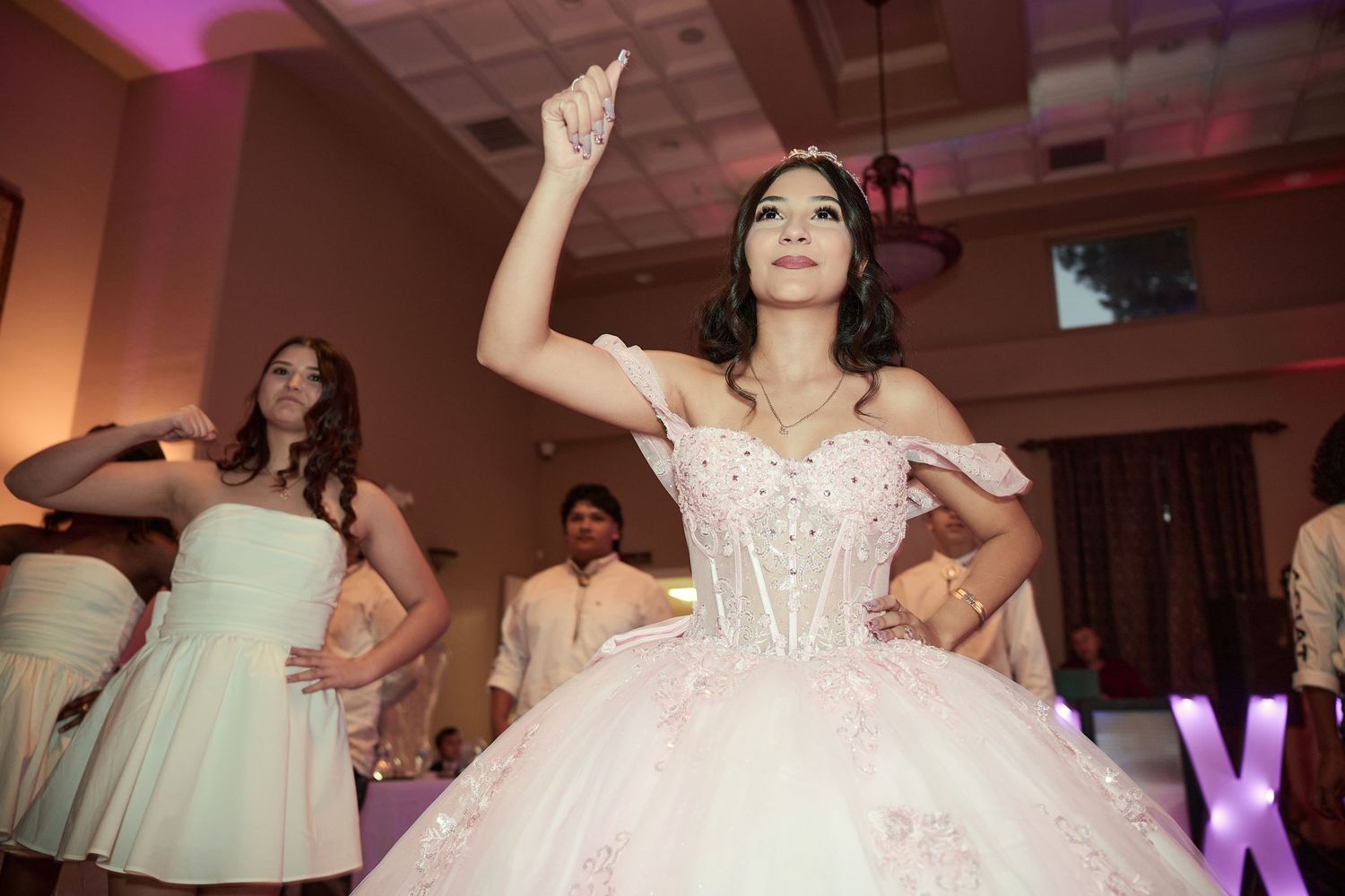 A group of dancers in formal attire perform at a wedding reception with pink and purple lighting.