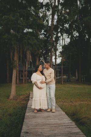 Couple in white and beige outfits walking together on a wooden boardwalk surrounded by trees.