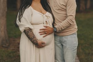 Expectant couple in casual attire embracing outdoors with focus on pregnant belly in white dress.
