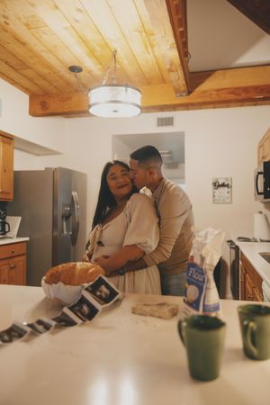 A couple shares an intimate moment in a kitchen with wooden ceiling beams and a green mug on the counter.