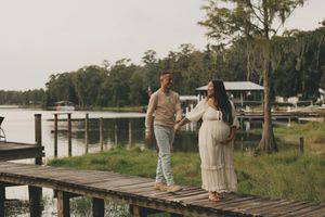 A couple walks along a rustic wooden dock surrounded by marsh grass and trees during golden hour.