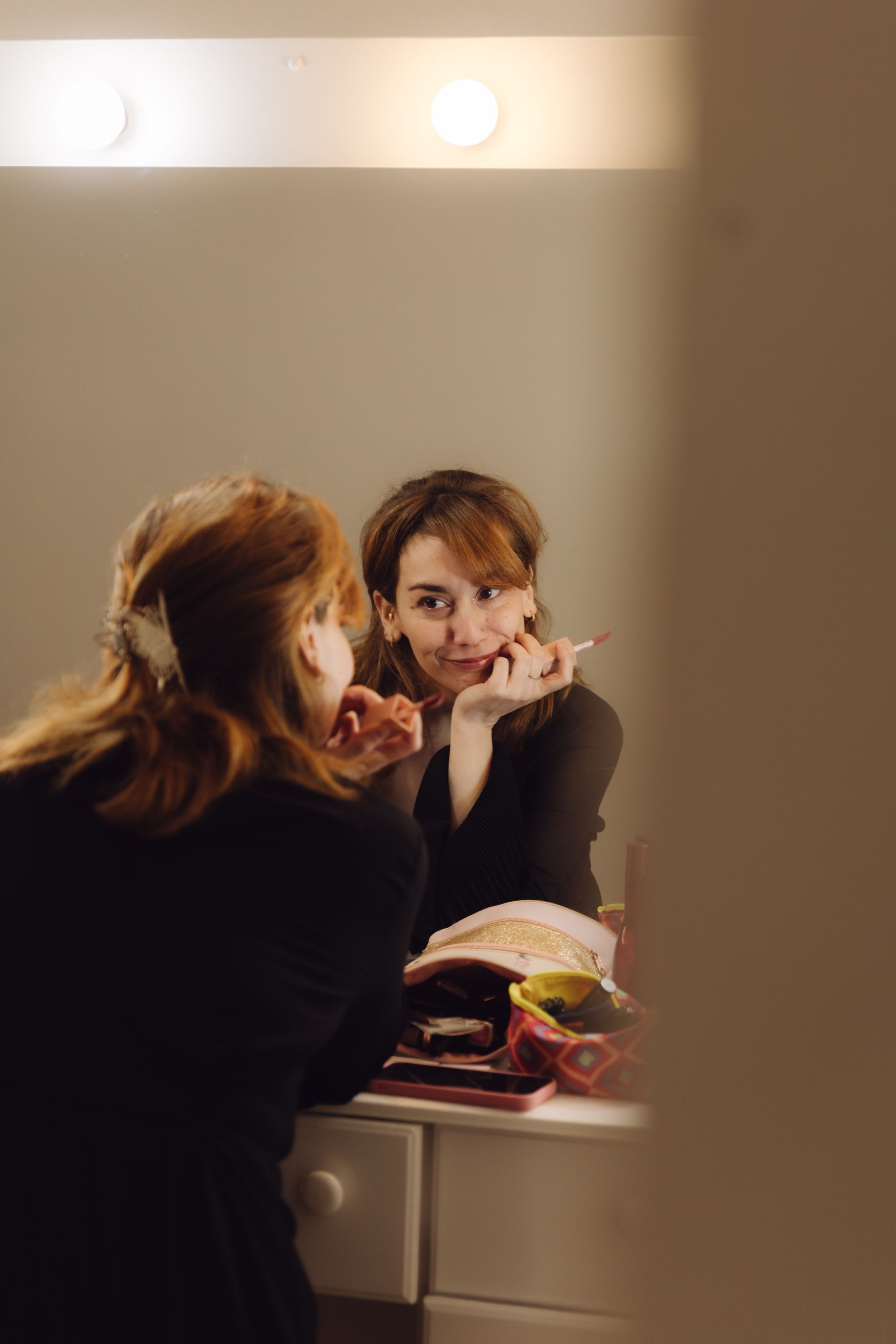 Young woman applying makeup while gazing into a illuminated vanity mirror in a warmly lit room.