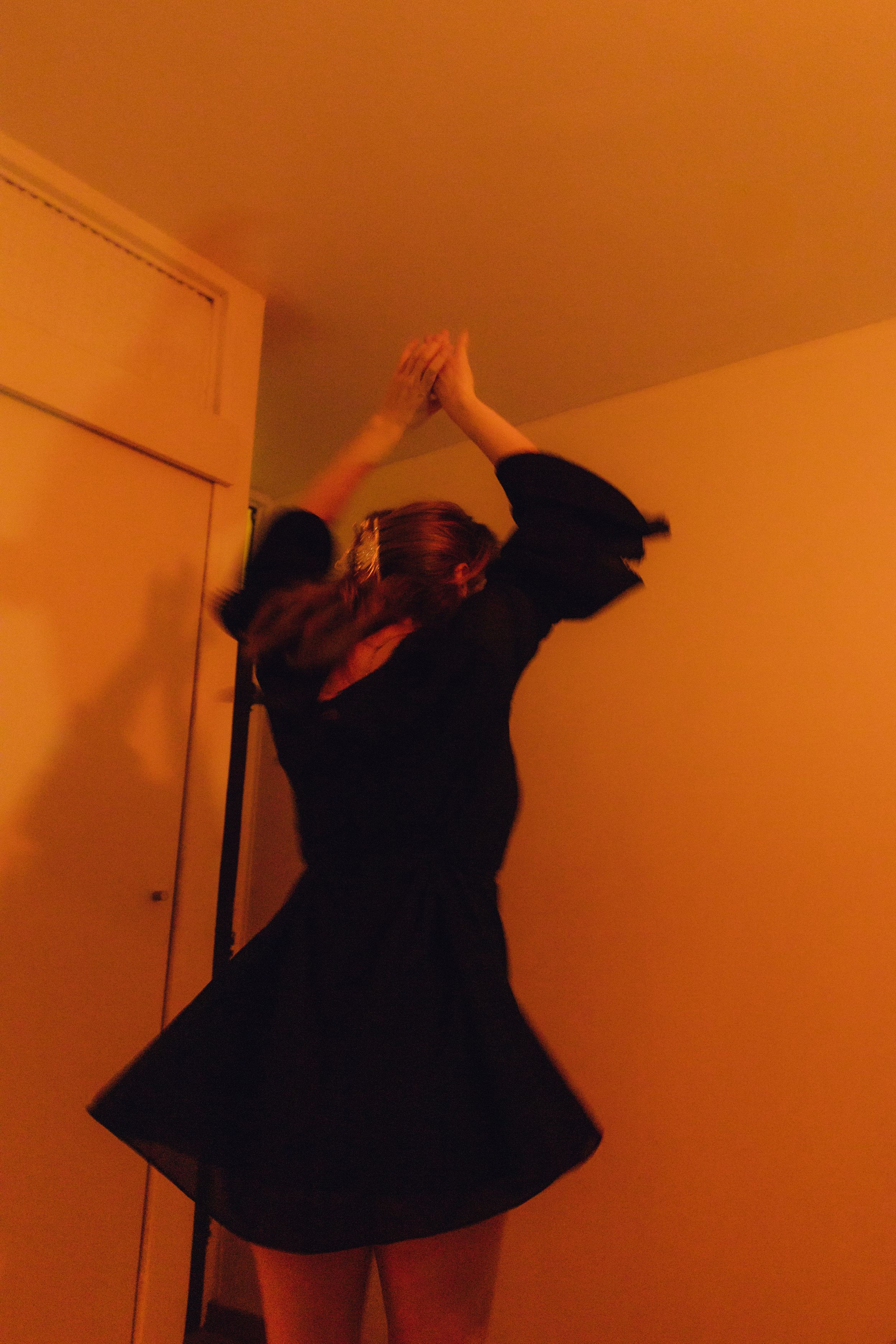 Woman in black dress reaching upward in an orange-lit room, captured with motion blur effect.